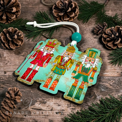 A three-piece Christmas nutcracker ornament set with a distressed turquoise background and colorful illustrations of three nutcrackers, displayed on a rustic wooden surface with pine needles and pinecones. A white hanging string with a turquoise bead is attached to the top.
