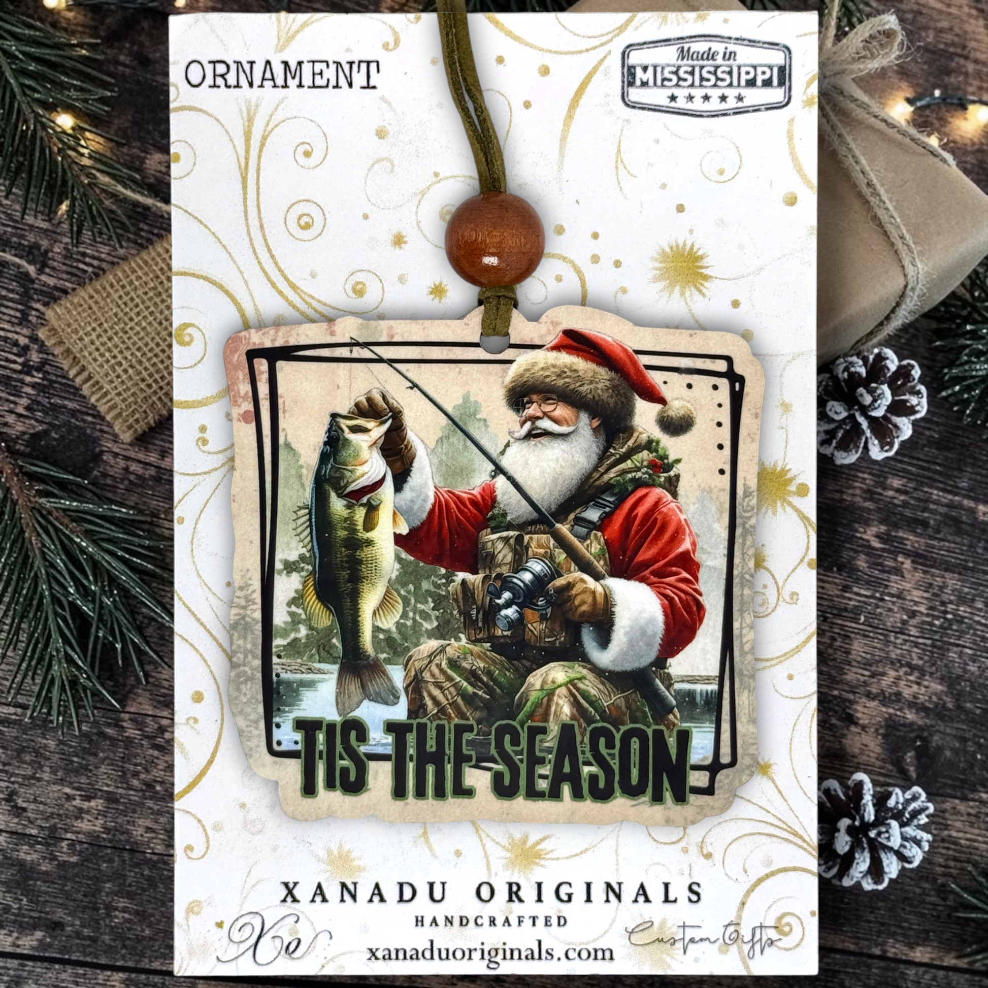 A Christmas ornament on a card featuring a handcrafted ornament with a Santa Claus fishing illustration and the text 'Tis the Season.' The ornament hangs from a brown cord with a wooden bead, displayed on a branded card with festive pine branches and string lights around it.