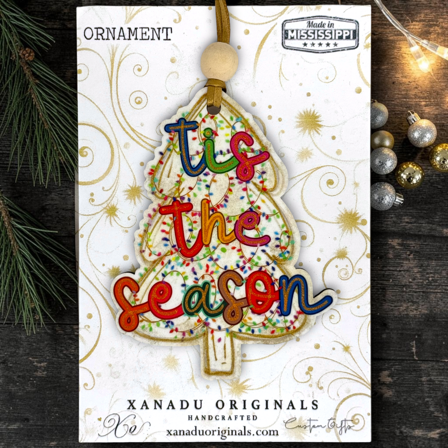 A Christmas ornament shaped like a tree with the phrase 'tis the season' in colorful raised letters, displayed on its branded retail card. The card reads 'ORNAMENT' at top and 'XANADU ORIGINALS HANDCRAFTED xanaduoriginals.