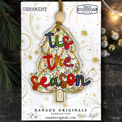 A Christmas ornament shaped like a tree with the phrase 'tis the season' in colorful raised letters, displayed on its branded retail card. The card reads 'ORNAMENT' at top and 'XANADU ORIGINALS HANDCRAFTED xanaduoriginals.