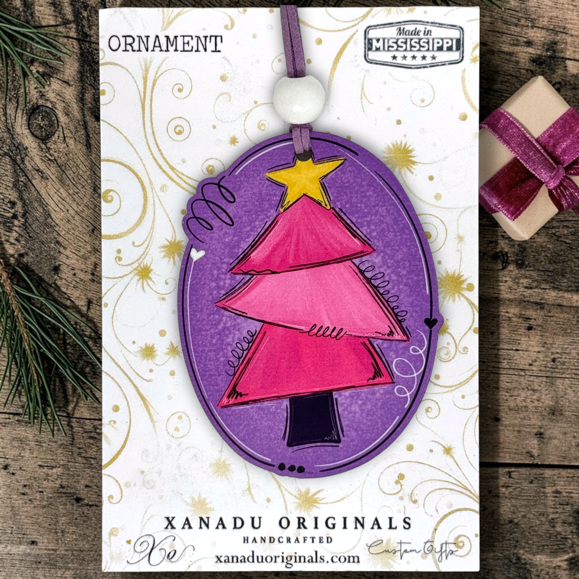 A Christmas ornament on a card featuring a handcrafted oval ornament with a pink tree design, purple border, yellow star, and white bead, displayed on branded Xanadu Originals cardstock over a rustic wood surface with pine and a small wrapped gift.