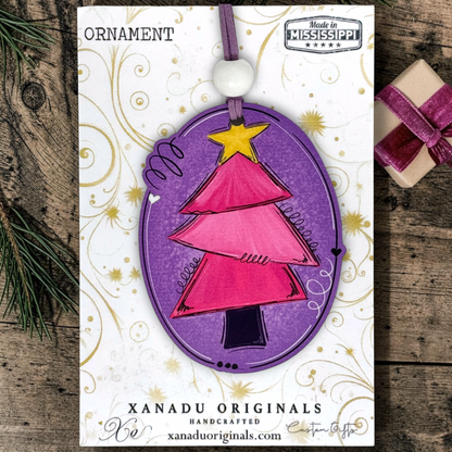 A Christmas ornament on a card featuring a handcrafted oval ornament with a pink tree design, purple border, yellow star, and white bead, displayed on branded Xanadu Originals cardstock over a rustic wood surface with pine and a small wrapped gift.