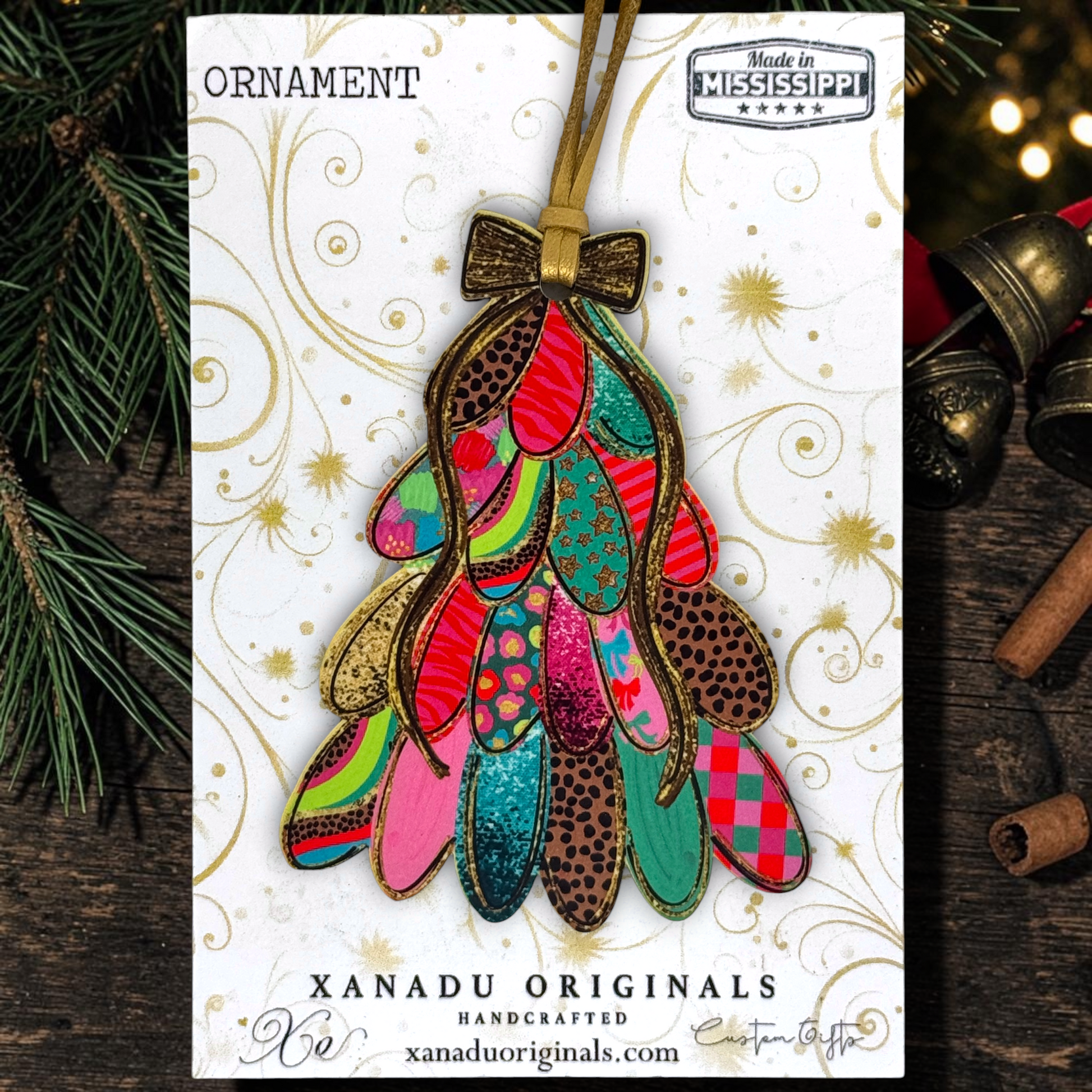 A Christmas ornament shaped like a mistletoe tree, featuring colorful patterned feathers in pink, green, blue, red, and brown, hanging from a gold ribbon. The ornament is displayed on its white printed backing card, which reads 'ORNAMENT' and 'XANADU ORIGINALS HANDCRAFTED', with pine branches and brass bells in the background.