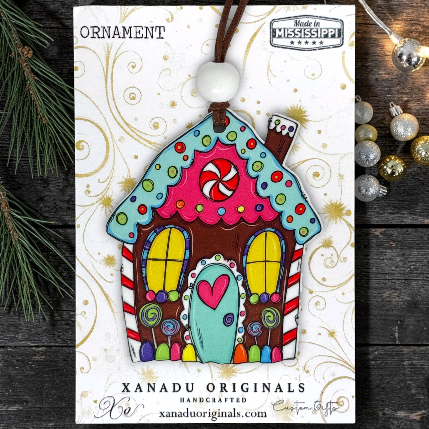 A colorful gingerbread house ornament carded on a white printed display card. The ornament has a brown top loop, a white bead, and is decorated with bright pink, teal, yellow, red, and brown icing-style patterns.
