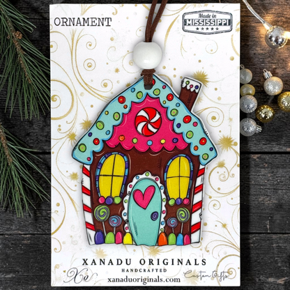 A colorful gingerbread house ornament carded on a white printed display card. The ornament has a brown top loop, a white bead, and is decorated with bright pink, teal, yellow, red, and brown icing-style patterns.