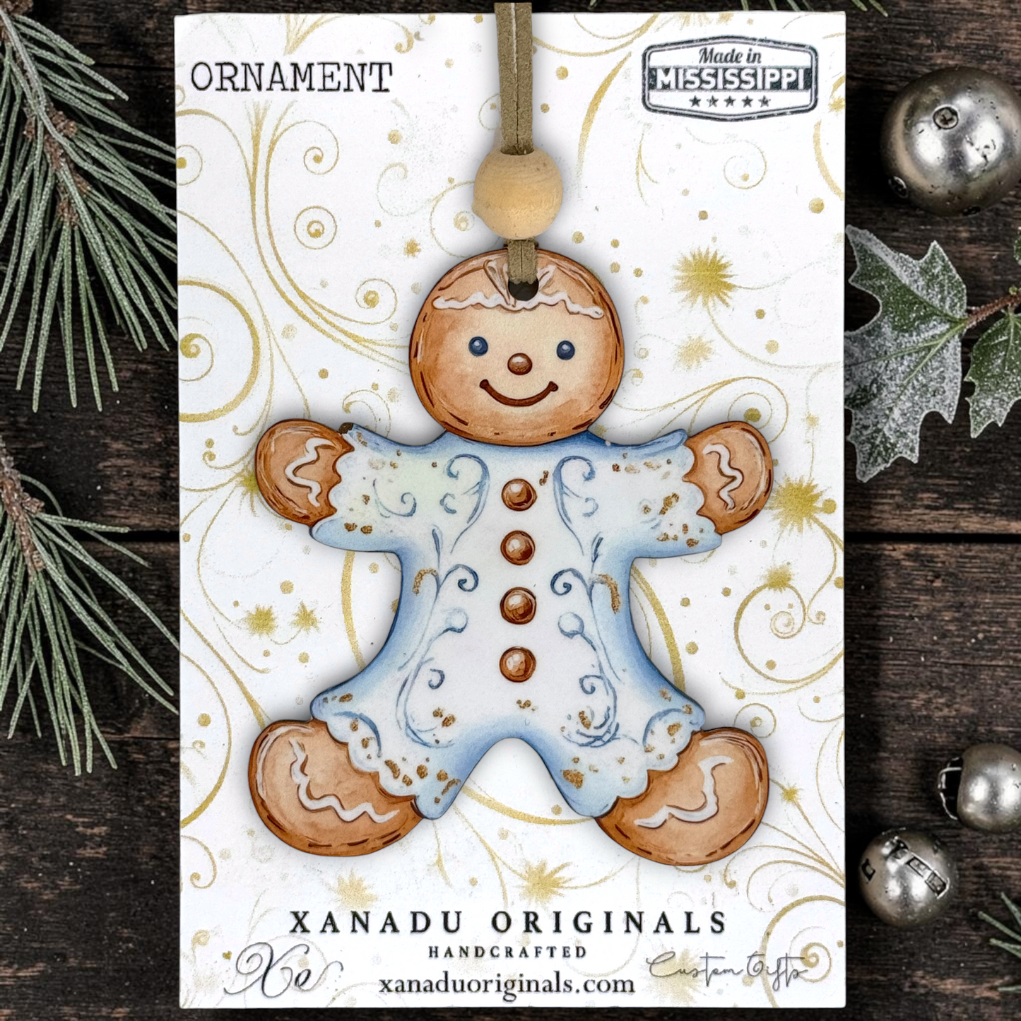 A gingerbread man ornament on a printed display card, photographed flat on a dark wood surface with pine branches and silver ornaments around it. The card shows the text 'ORNAMENT,' 'Made in Mississippi,' and 'XANADU ORIGINALS HANDCRAFTED xanaduoriginals.