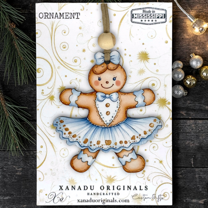 A handcrafted gingerbread doll Christmas ornament by Xanadu Originals, displayed on its white printed backing card. The ornament features a gingerbread figure dressed in a light blue tutu and bow tie, with a wooden bead hanging loop.