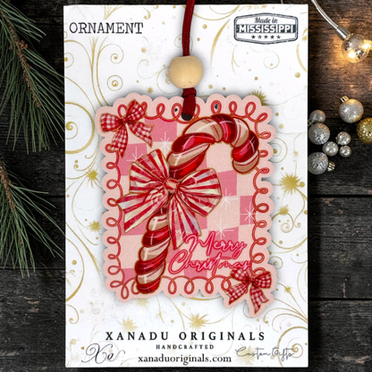 A Christmas ornament card from Xanadu Originals featuring a red and white striped candy cane ornament with a wooden bead hanging cord. The card has gold swirls and the words 'ORNAMENT' and 'Made in Mississippi,' and is photographed on a dark wood surface with pine branches and small ornaments around it.