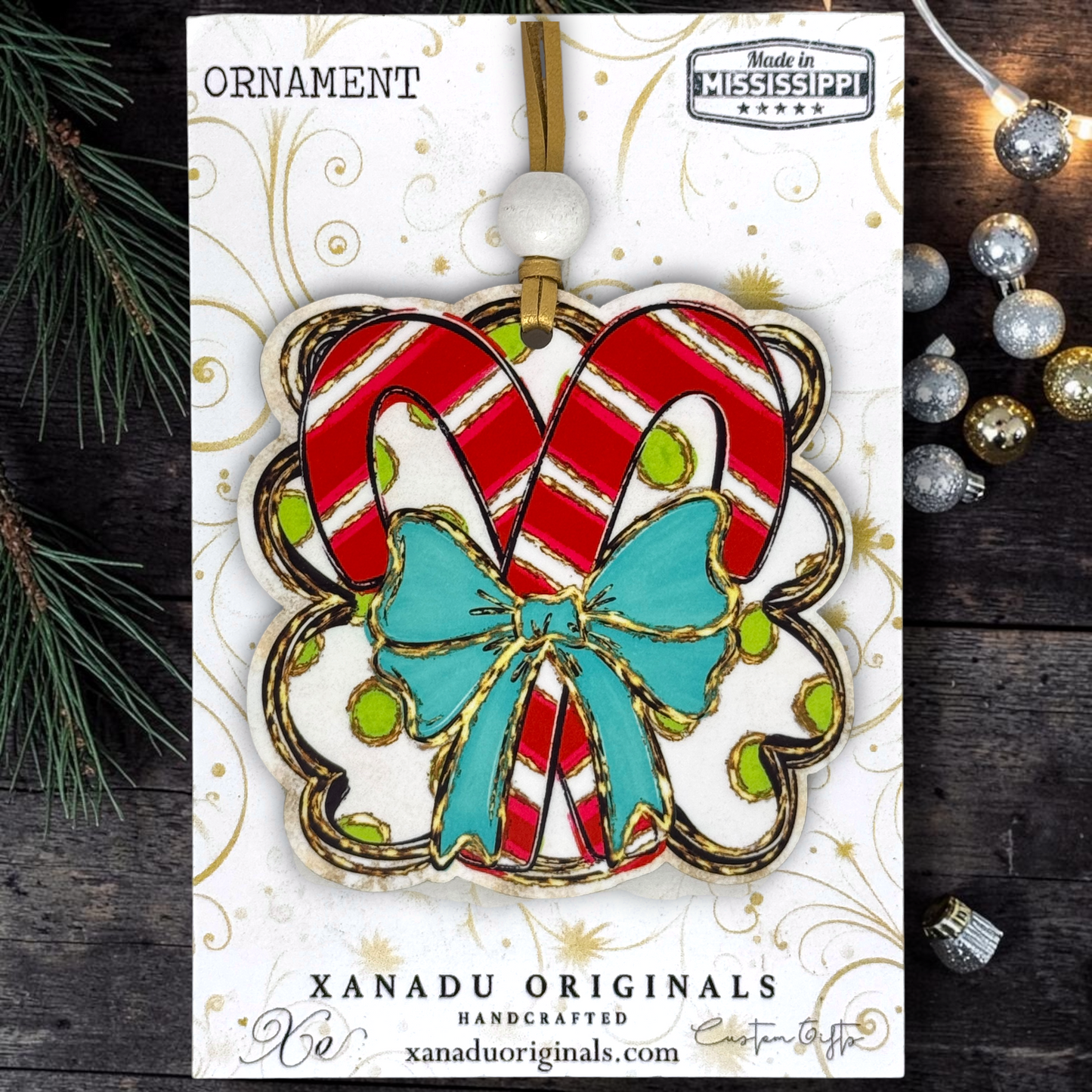A Christmas ornament card featuring a candy cane-shaped ornament with red and white stripes, a turquoise bow, and gold glitter details, displayed on a branded card with holiday props.
