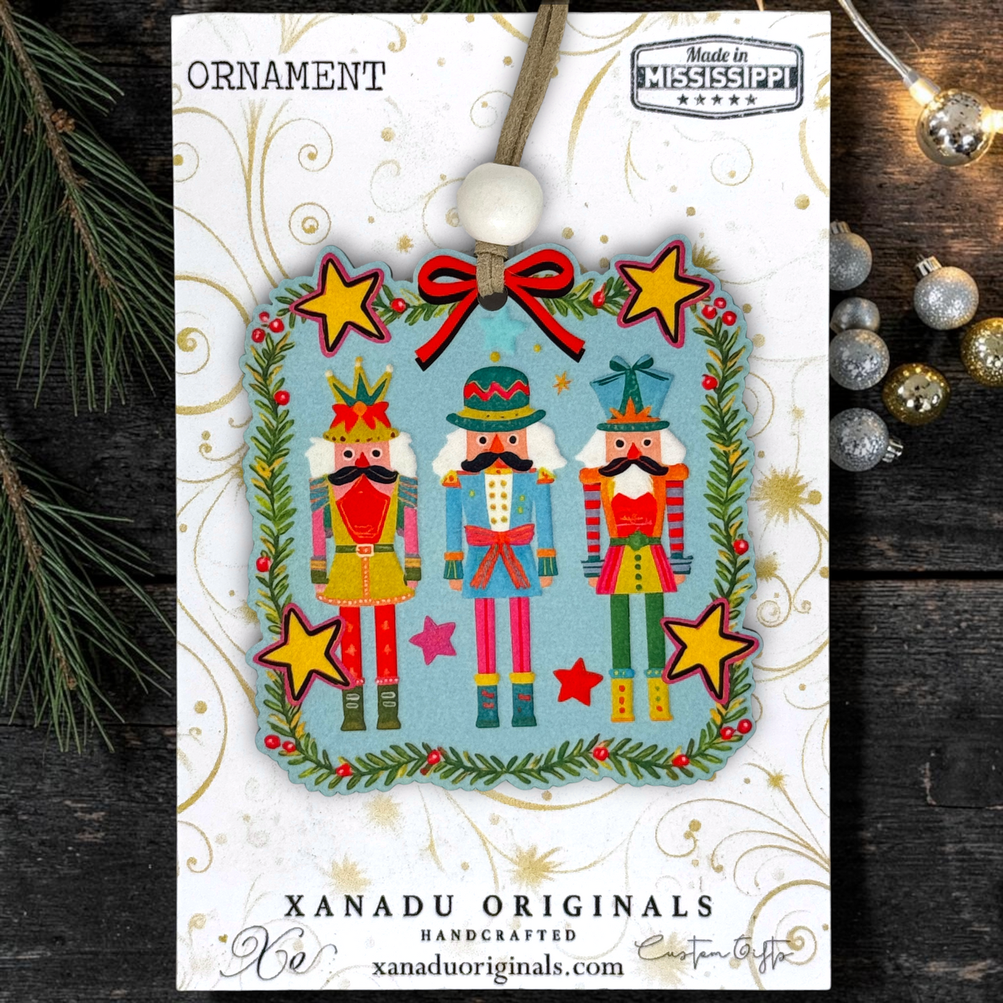 A Christmas ornament card featuring three nutcracker figures framed by a green wreath with red berries and yellow stars, hanging from a tan cord with a white bead. The card is branded XANADU ORIGINALS and photographed on a dark wood surface with pine branches and silver ornaments around it.
