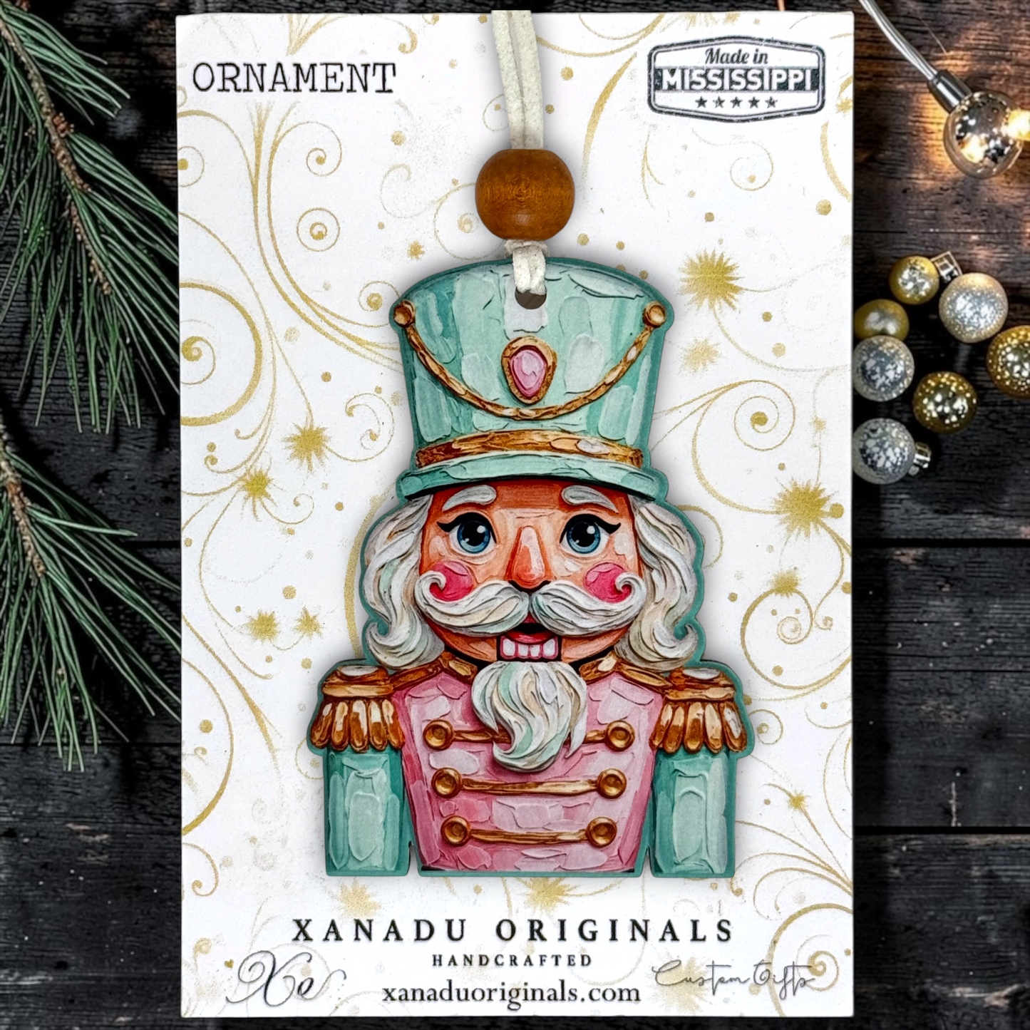 A handcrafted Christmas ornament shaped like a nutcracker soldier, displayed on its branded backing card. The ornament features a mint green hat, pink coat, white beard, and gold accents, with a wooden bead at the top.