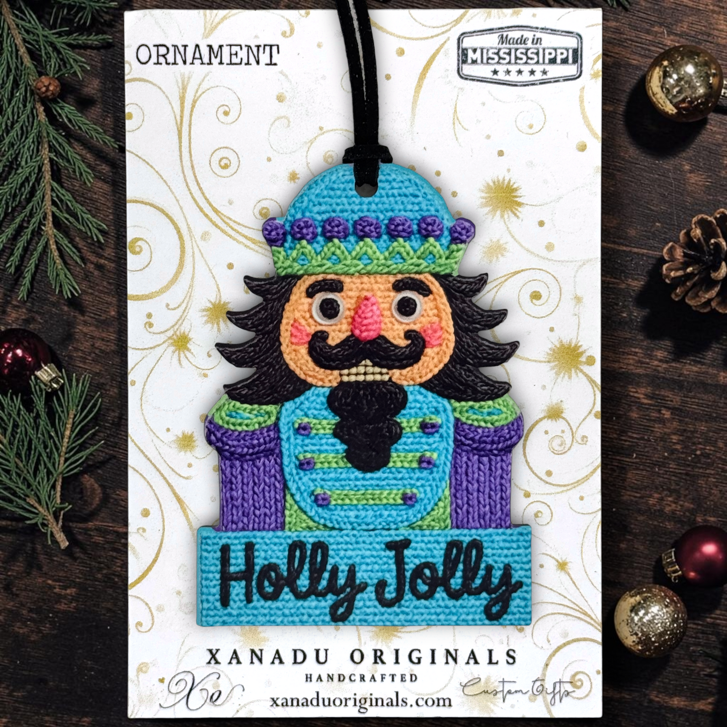 A handcrafted embroidered Christmas nutcracker ornament carded on a white printed display card. The ornament features a colorful embroidered nutcracker with a blue hat, purple coat, black beard, and pink cheeks, with 'Holly Jolly' stitched on a blue banner.