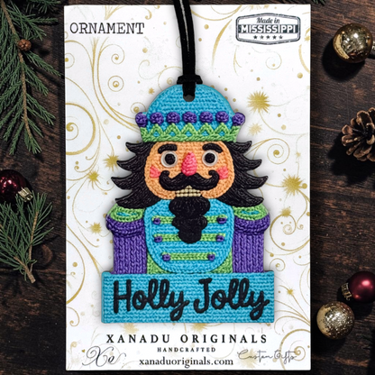 A handcrafted embroidered Christmas nutcracker ornament carded on a white printed display card. The ornament features a colorful embroidered nutcracker with a blue hat, purple coat, black beard, and pink cheeks, with 'Holly Jolly' stitched on a blue banner.