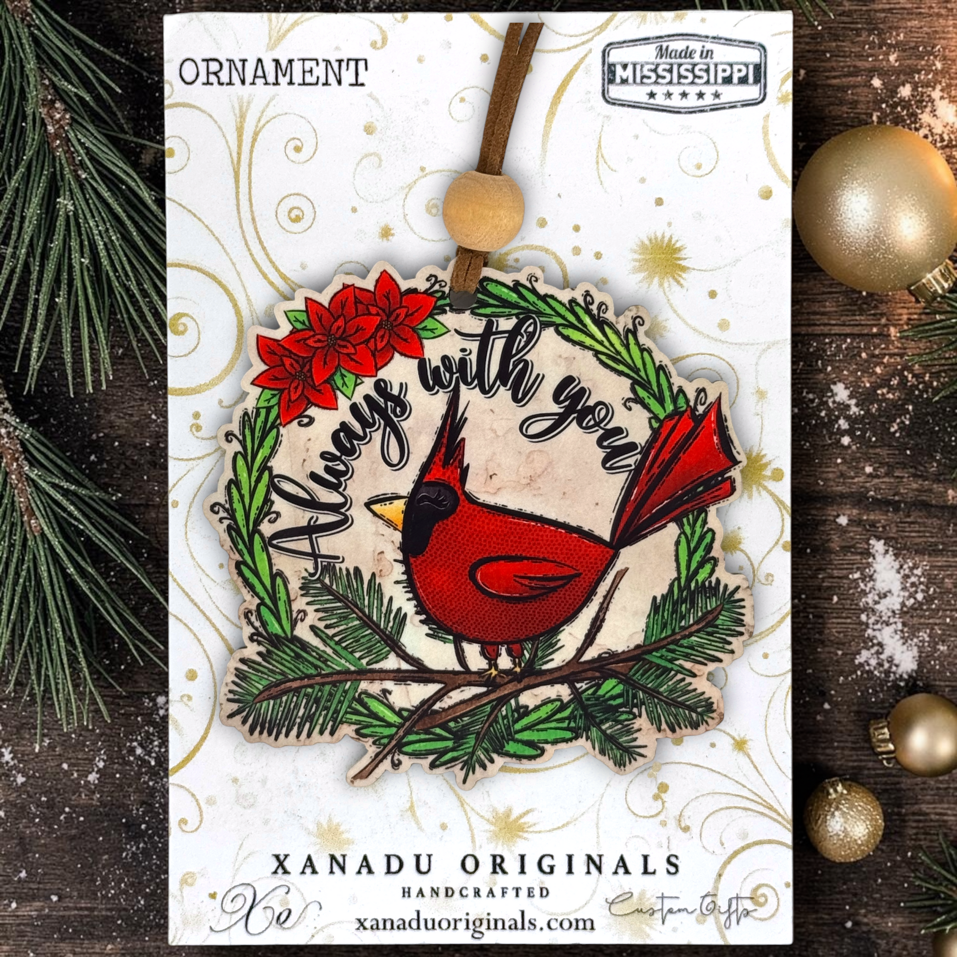 A Christmas ornament card featuring a red cardinal perched on a green wreath with the phrase “Always with you,” shown on a branded XANADU ORIGINALS card with holiday decorations around it.
