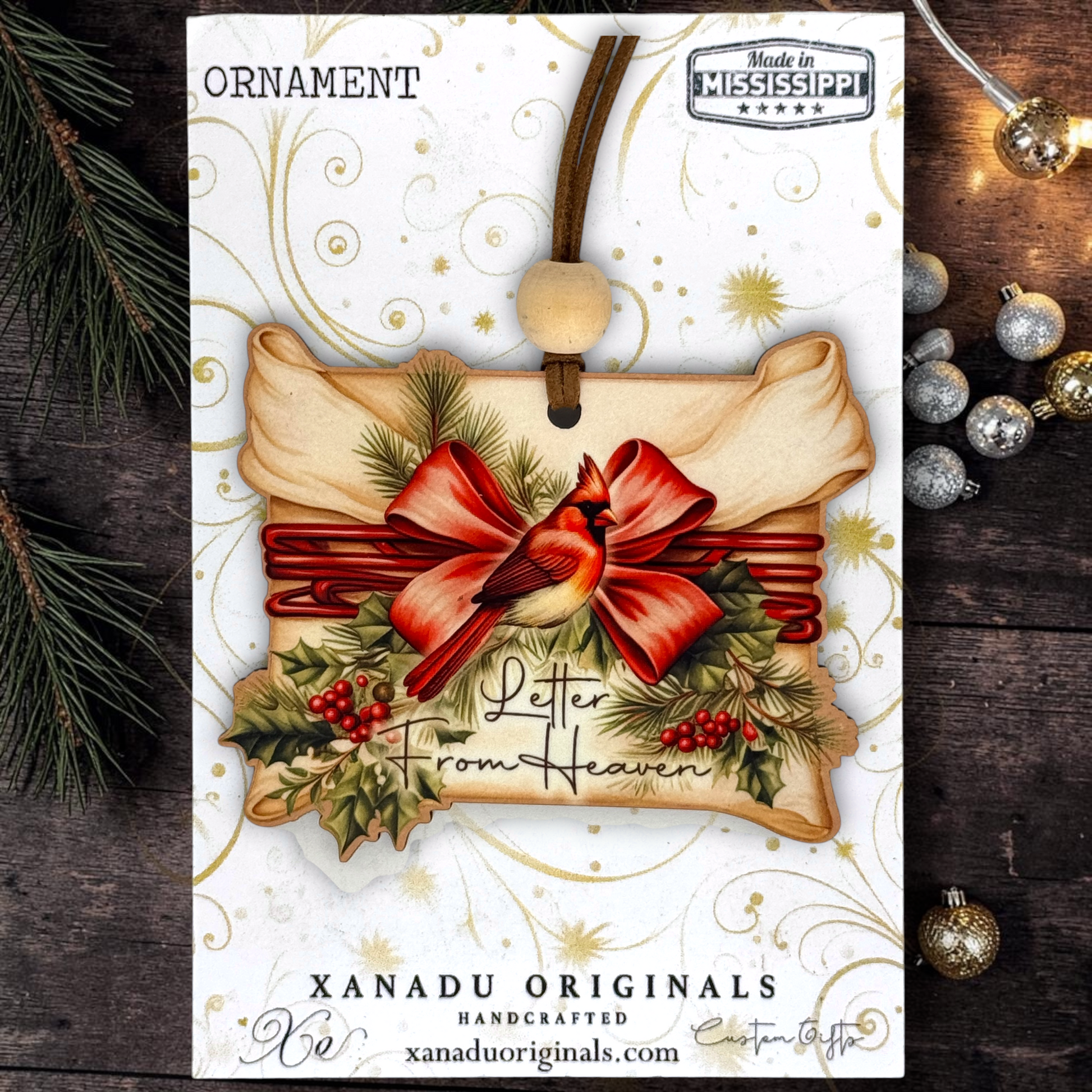 A Christmas ornament card featuring a handcrafted ornament design with a red cardinal, red bow, green holly leaves, and the words 'Letter From Heaven.' The card is branded 'XANADU ORIGINALS' and includes a 'Made in Mississippi' badge, shown on a dark wood background with holiday ornaments and evergreen branches.