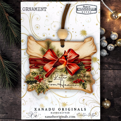A Christmas ornament card featuring a handcrafted ornament design with a red cardinal, red bow, green holly leaves, and the words 'Letter From Heaven.' The card is branded 'XANADU ORIGINALS' and includes a 'Made in Mississippi' badge, shown on a dark wood background with holiday ornaments and evergreen branches.