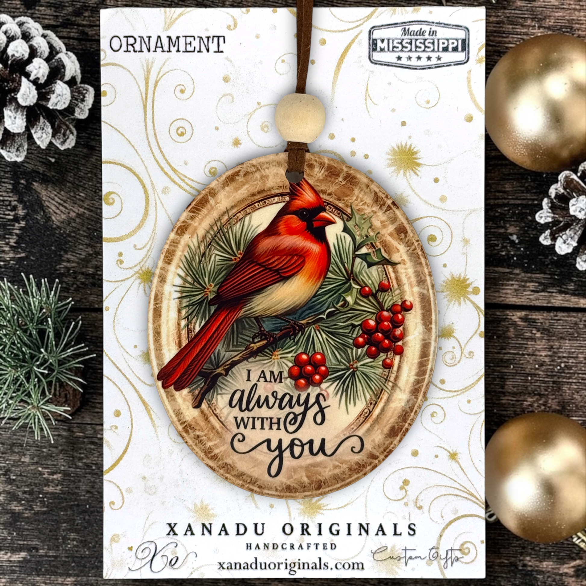 A oval wooden Christmas ornament with a red cardinal perched among pine branches and red berries, with the phrase “I am always with you” printed below. The ornament hangs from a brown cord and is displayed on a branded card, surrounded by pinecones and gold ornaments on a dark wood surface.