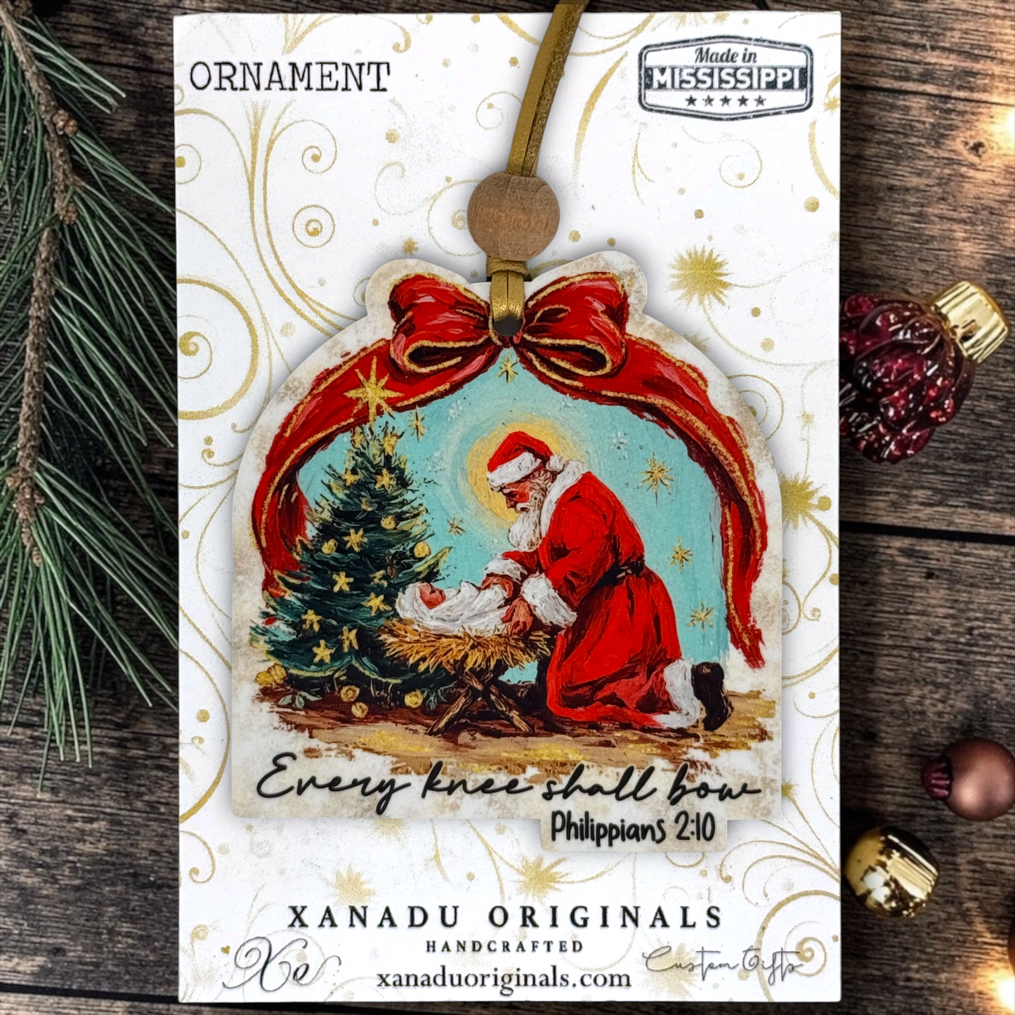 A Christmas ornament card featuring a handcrafted ornament with an illustration of Saint Christopher and baby Jesus under a red bow, with text reading 'ORNAMENT' and 'XANADU ORIGINALS HANDCRAFTED'.