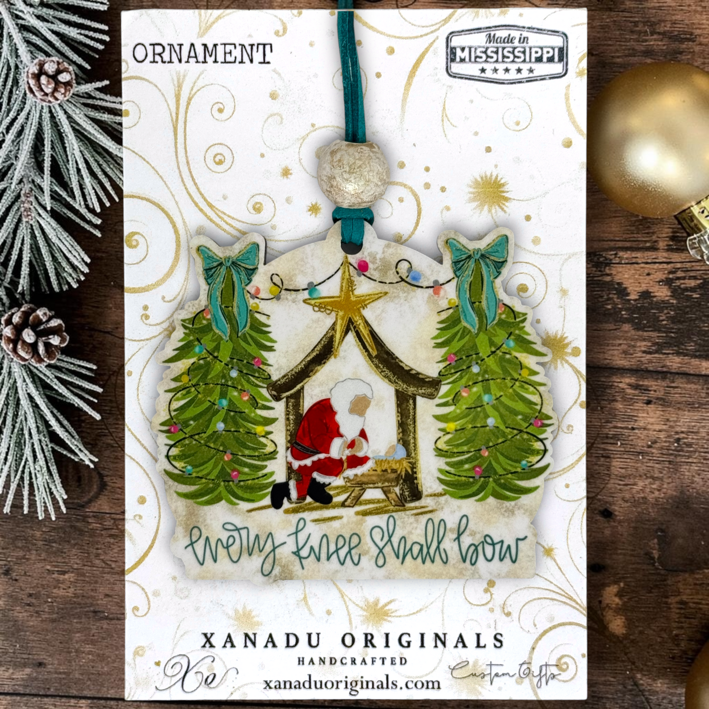 Handcrafted Christmas ornament package by Xanadu Originals, featuring a nativity scene ornament with two green trees, a gold star, and the phrase 'Every Tree Shall Bow,' displayed on a rustic wooden surface with holiday decorations.