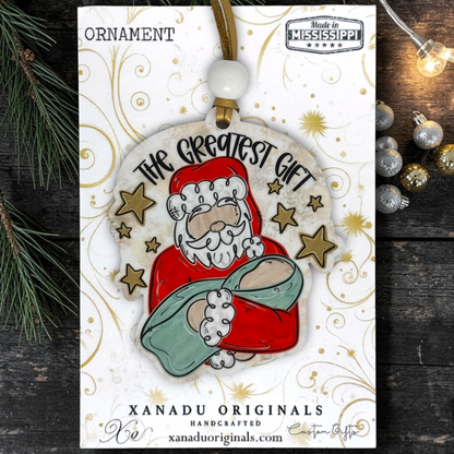 Handcrafted Christmas ornament on its display card, featuring Santa Claus holding a baby wrapped in a green blanket, with the text 'The Greatest Gift'.