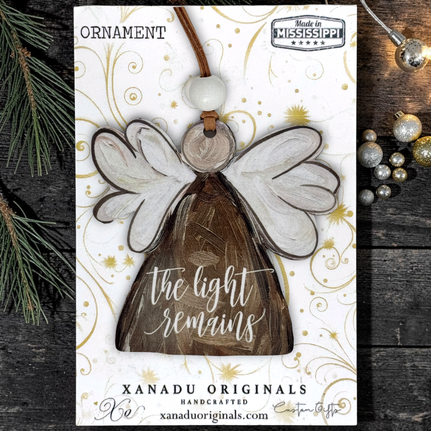 Handcrafted wooden angel ornament on a white display card with gold swirls, photographed on a dark wood surface with pine branches and silver ornaments around it. The card reads 'ORNAMENT' and 'XANADU ORIGINALS HANDCRAFTED xanaduoriginals.