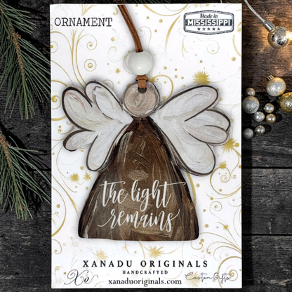 Handcrafted wooden angel ornament on a white display card with gold swirls, photographed on a dark wood surface with pine branches and silver ornaments around it. The card reads 'ORNAMENT' and 'XANADU ORIGINALS HANDCRAFTED xanaduoriginals.