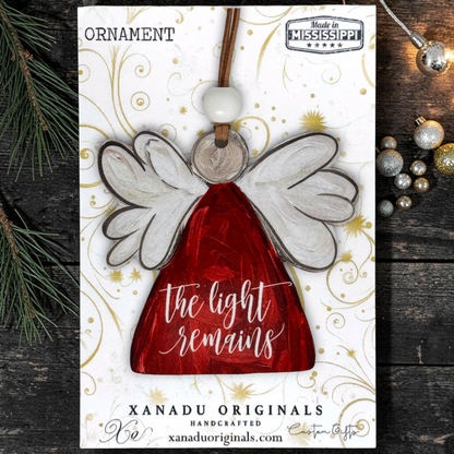 Handcrafted wooden angel ornament by Xanadu Originals, featuring a red dress, white wings, and the phrase 'the light remains' on a white card with gold swirls, styled on a dark wood background with pine branches and holiday ornaments.