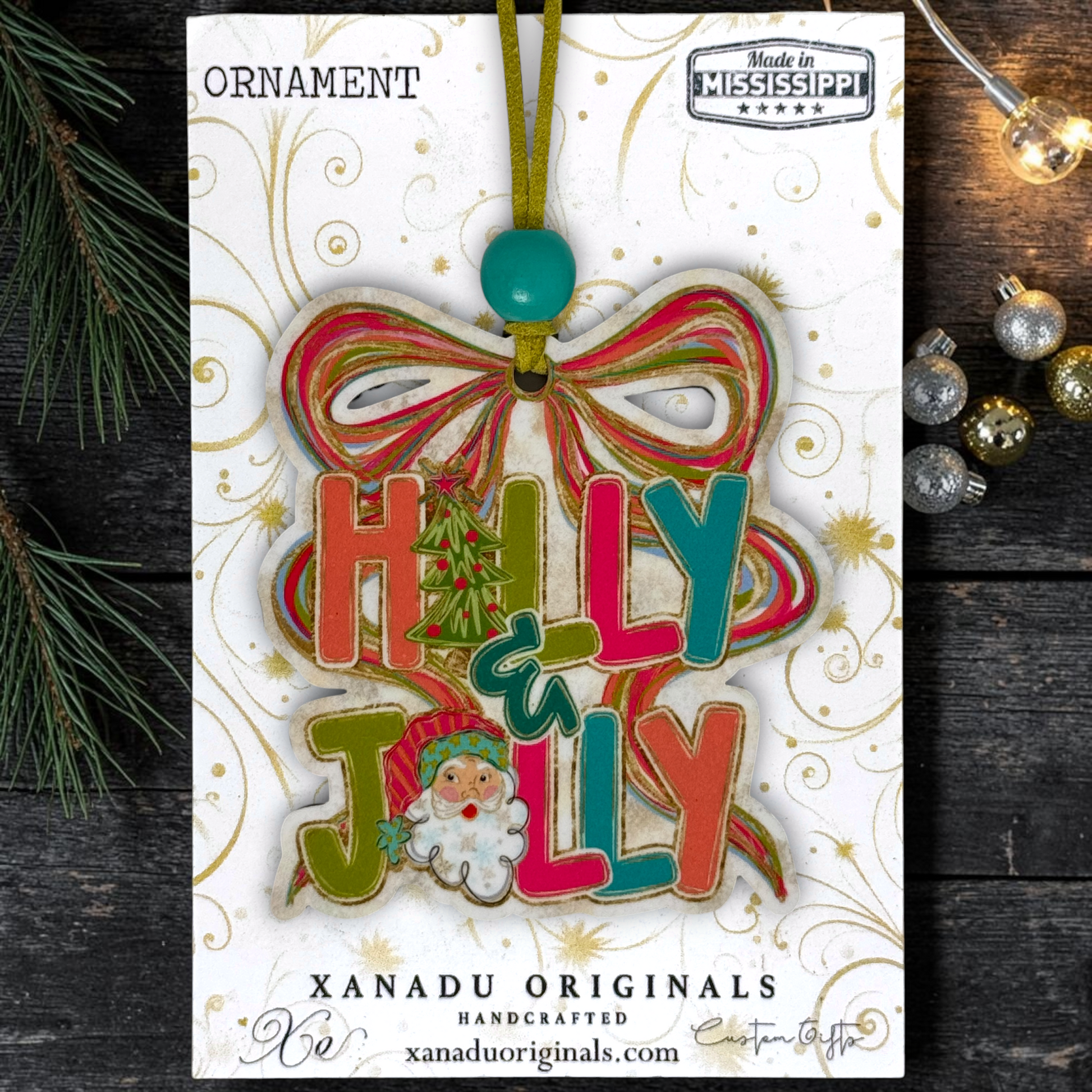 A festive Christmas ornament on a card featuring a colorful 'Holly Jolly' ornament with a green hanging loop and turquoise bead, shown on a dark wood background with pine branches and holiday ornaments.