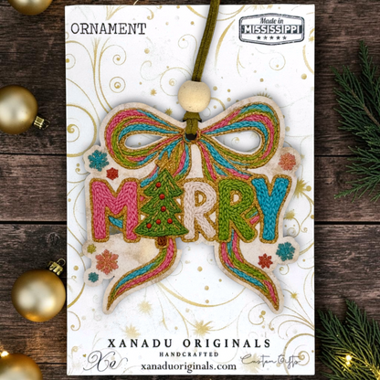 A festive embroidered Christmas ornament card featuring a bow and the word “MERRY” in colorful thread, with “ORNAMENT” at the top and “XANADU ORIGINALS” at the bottom. The card is photographed on a rustic dark wood surface with gold ornaments and evergreen branches around it.