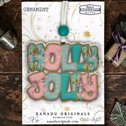 A handcrafted Christmas ornament on a card featuring the words “HOLLY JOLLY” in pink and turquoise icing with gold glitter details, hanging from a white cord with a turquoise bead. The card is branded XANADU ORIGINALS and photographed on a rustic dark wood surface with decorative holiday props.