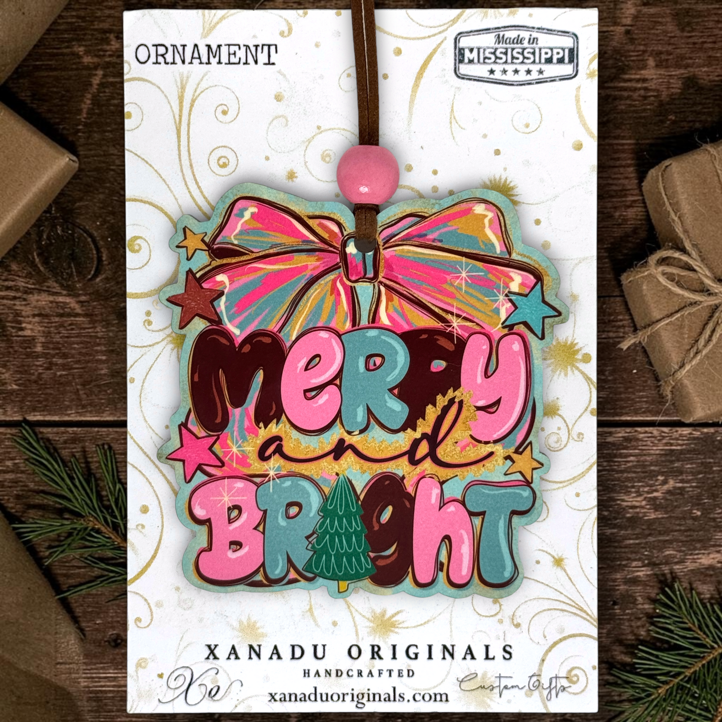 A colorful Christmas ornament card featuring a bow and the words “Merry and Bright” in layered pink, teal, brown, and gold lettering, with a pink bead at the top. The card is photographed on a dark wood surface with wrapped gifts and pine branches around it.