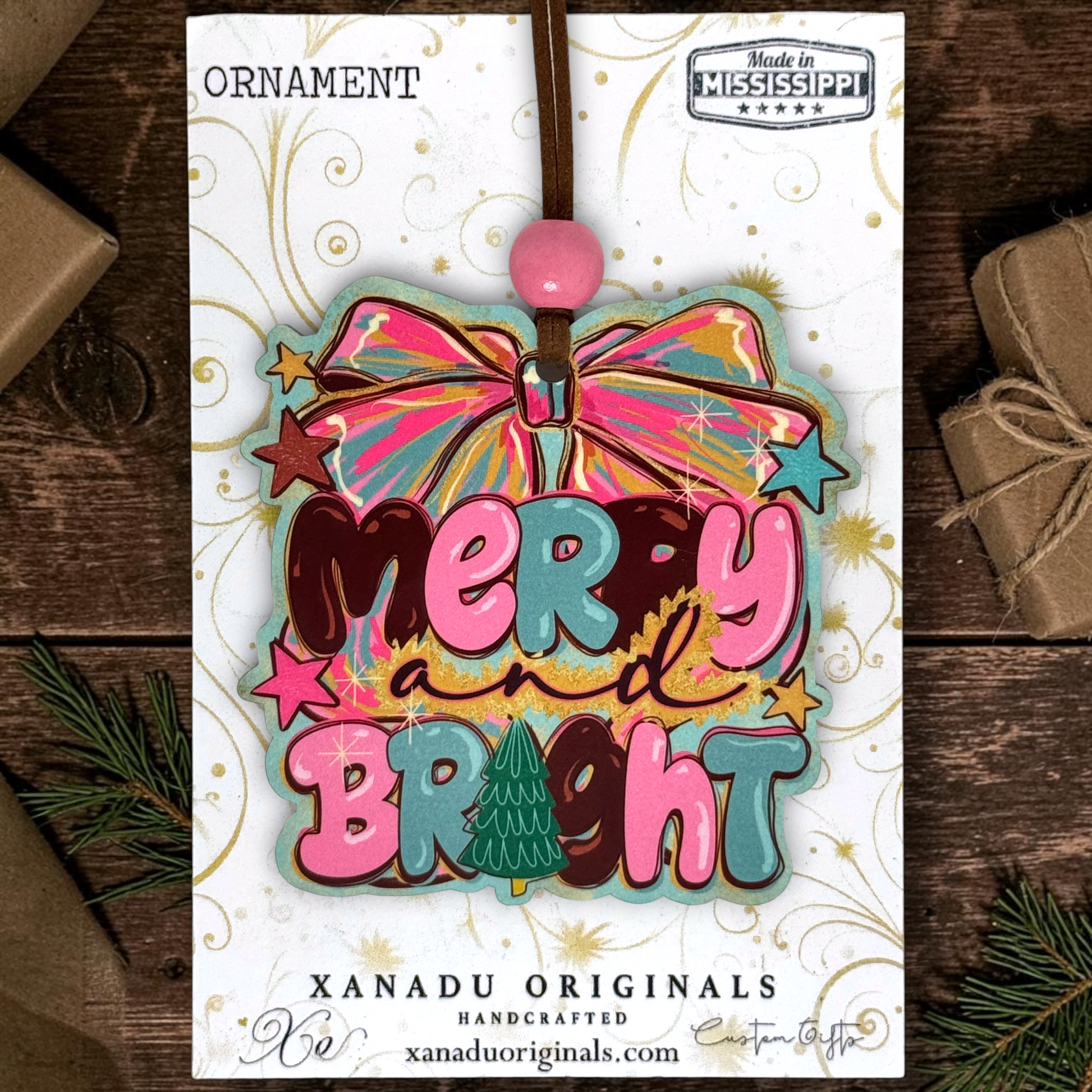 A colorful Christmas ornament card featuring a bow and the words “Merry and Bright” in layered pink, teal, brown, and gold lettering, with a pink bead at the top. The card is photographed on a dark wood surface with wrapped gifts and pine branches around it.