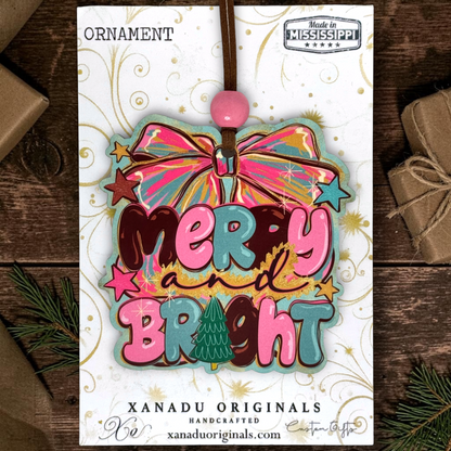 A colorful Christmas ornament card featuring a bow and the words “Merry and Bright” in layered pink, teal, brown, and gold lettering, with a pink bead at the top. The card is photographed on a dark wood surface with wrapped gifts and pine branches around it.