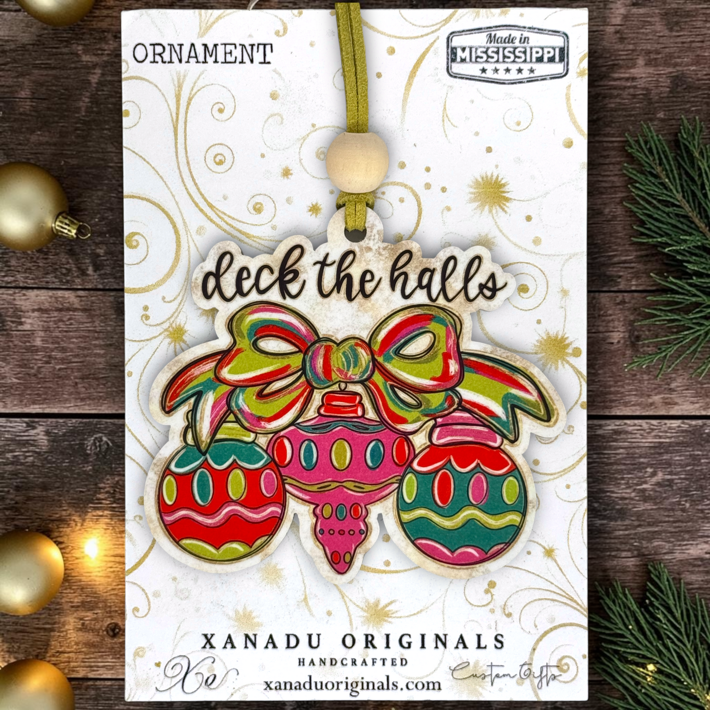 A Christmas ornament card featuring a colorful handcrafted ornament with three hanging ornaments and a bow, printed with the words 'ORNAMENT' and 'deck the halls,' from Xanadu Originals. The card is photographed on a rustic dark wood surface with gold ornaments and evergreen branches around it.