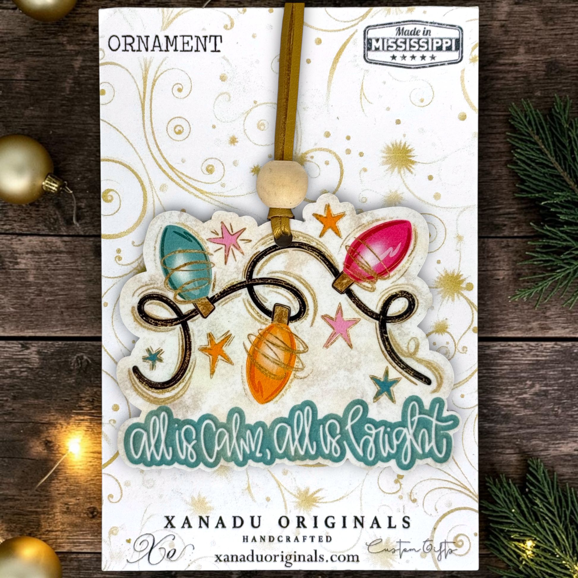 A Christmas ornament card featuring a handmade ornament with colorful light bulbs and the phrase 'all is calm, all is light.' The card is displayed on a rustic dark wood background with holiday lights and pine branches.