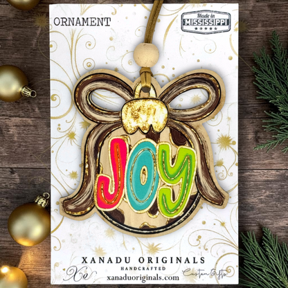 A wooden Christmas ornament shaped like a bauble with the word 'Joy' in colorful letters, displayed on its branded white backing card. The card is photographed on a rustic dark wood surface with gold ornaments and evergreen branches around it.