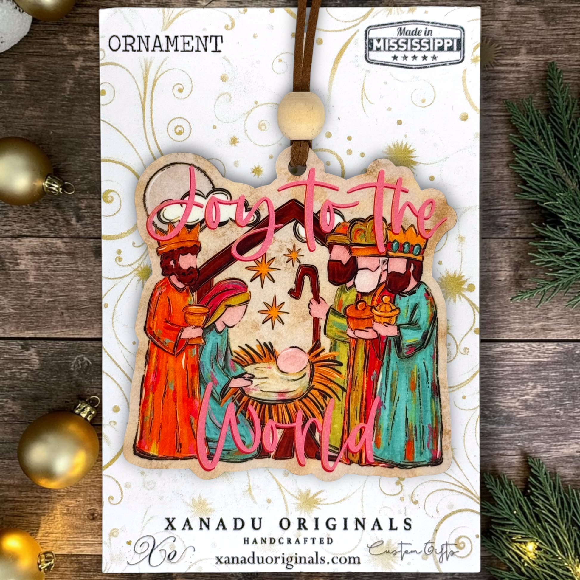 A Christmas ornament on a card featuring a handcrafted wooden nativity ornament with the text 'Joy to the World' in pink script. The ornament depicts the Holy Family, the baby Jesus, and the Three Wise Men.