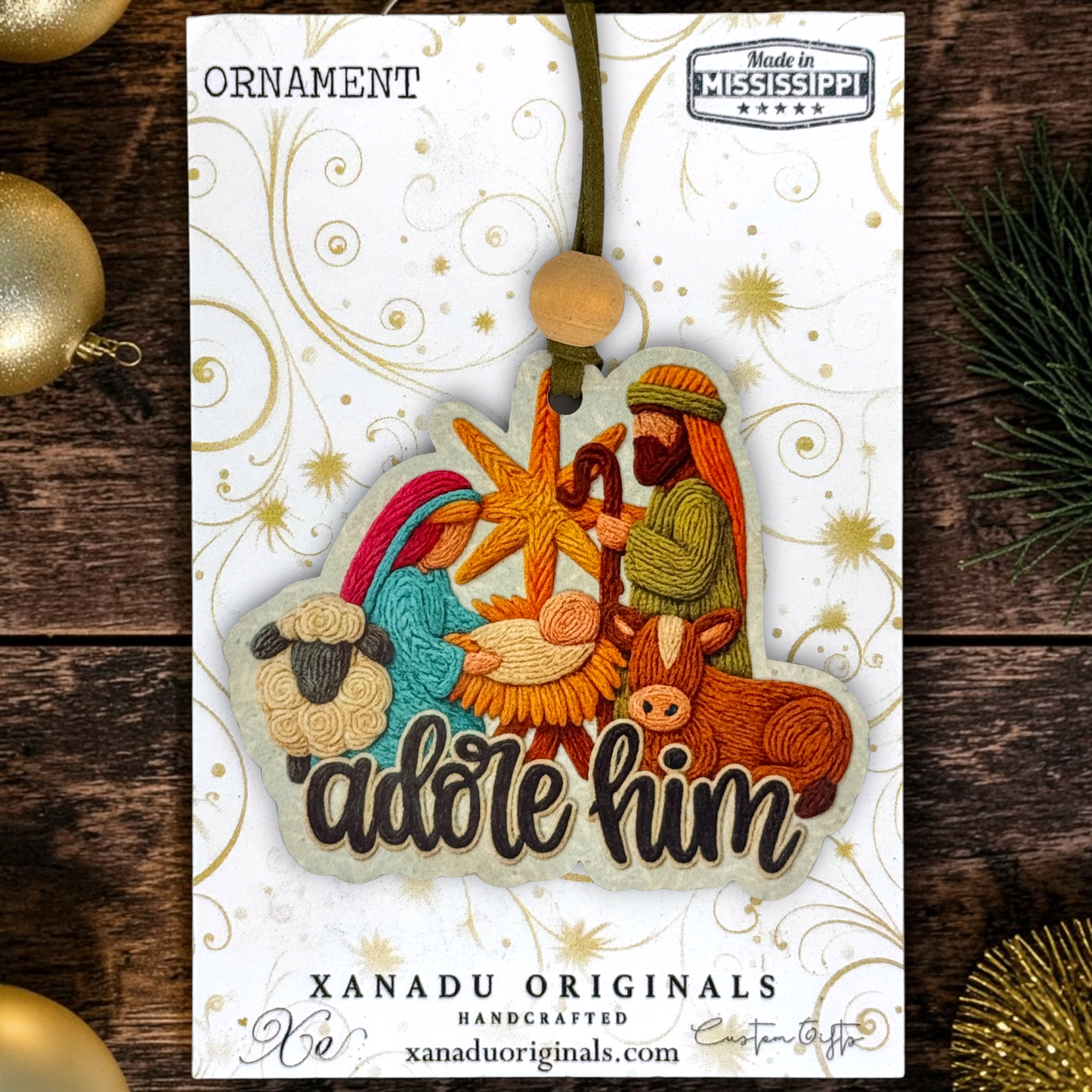 A handmade embroidered Christmas ornament featuring the Nativity scene with the words 'Adore Him' on a white display card. The card has gold swirls, a 'Made in Mississippi' badge, and 'XANADU ORIGINALS' branding.