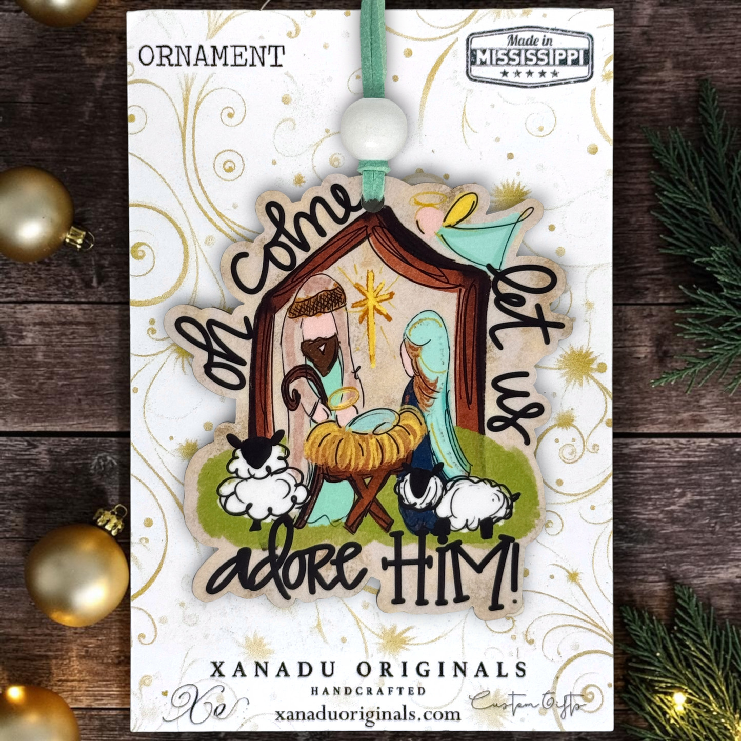 A handcrafted wooden Christmas ornament featuring a nativity scene with Mary, Joseph, baby Jesus in a manger, and sheep, surrounded by the words 'Oh come, let us adore HIM!'. The ornament is displayed on its white branded backing card, which reads 'XANADU ORIGINALS HANDCRAFTED' and 'Made in Mississippi'.