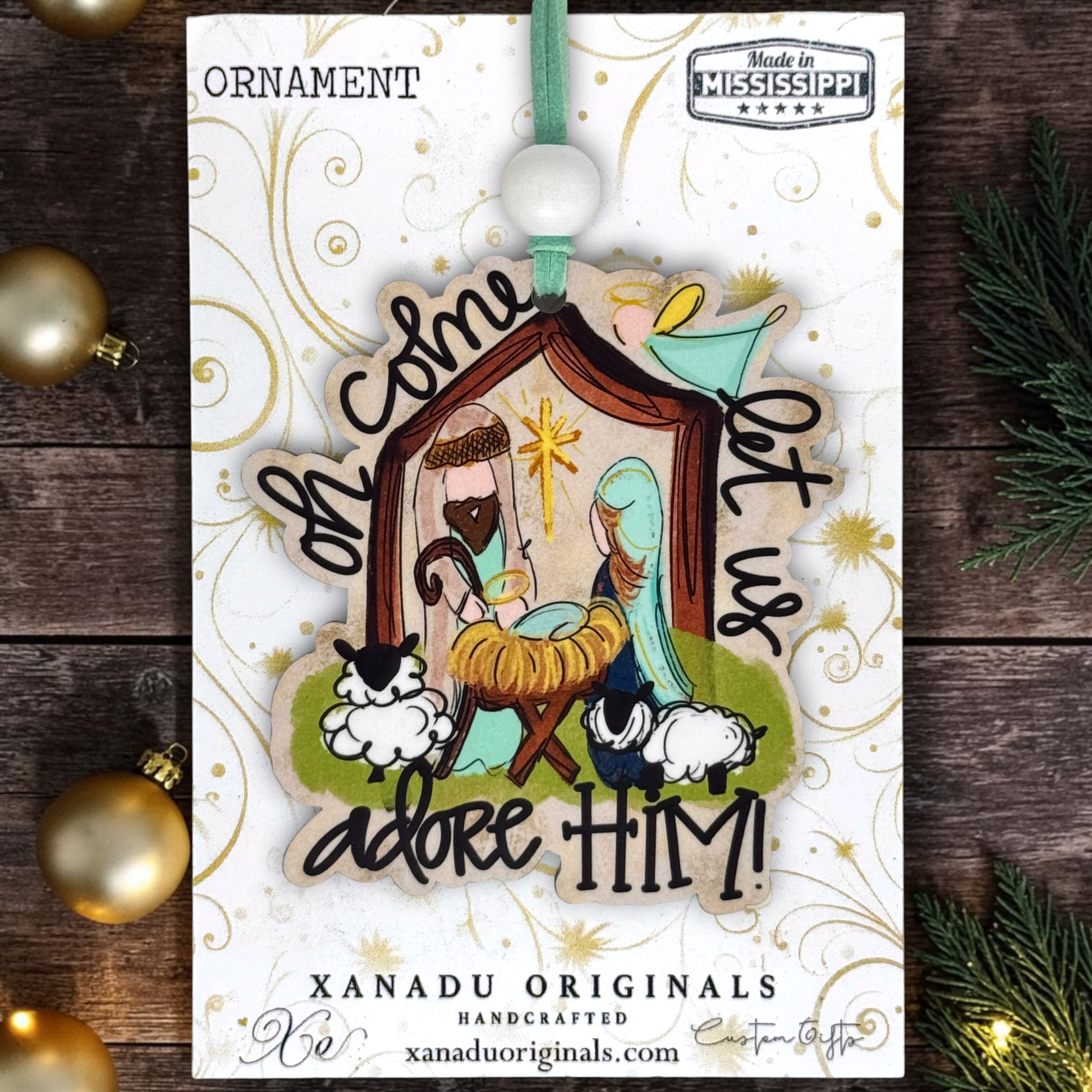 A handcrafted wooden Christmas ornament featuring a nativity scene with Mary, Joseph, baby Jesus in a manger, and sheep, surrounded by the words 'Oh come, let us adore HIM!'. The ornament is displayed on its white branded backing card, which reads 'XANADU ORIGINALS HANDCRAFTED' and 'Made in Mississippi'.