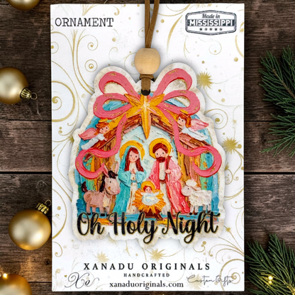 A handcrafted Christmas ornament package from Xanadu Originals, featuring a colorful wooden Nativity scene ornament with a pink bow, gold star, and the text 'Oh Holy Night' on a white card with gold swirls, set against a dark wood background with holiday decorations.