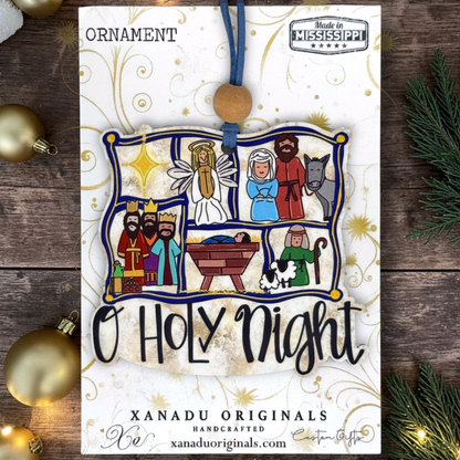 A handmade Christmas ornament featuring a colorful nativity scene illustration with the text 'O HOLY NIGHT' on a white card, surrounded by gold holiday decorations and evergreen branches on a dark wood background.