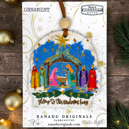 A handcrafted Christmas ornament on a card featuring a nativity scene illustration with colorful figures, a golden star, and the text 'Glory to the newborn king.' The ornament hangs from a brown cord with a round beige bead.