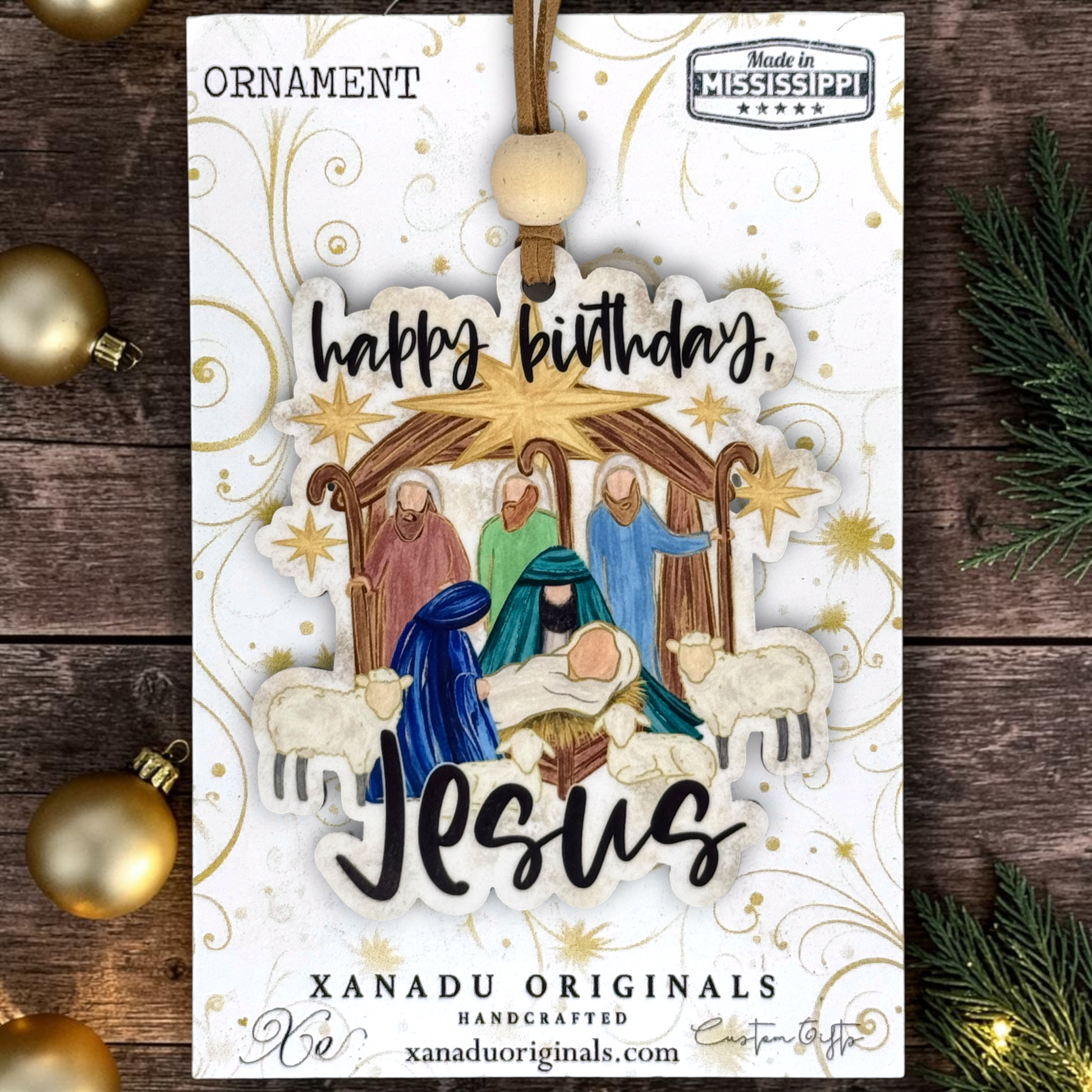A wooden Christmas ornament on a card featuring a handcrafted Nativity scene ornament with the words 'happy birthday, Jesus.' The ornament is displayed on a white card with gold decorative swirls, surrounded by gold baubles and evergreen branches on a dark wood surface.