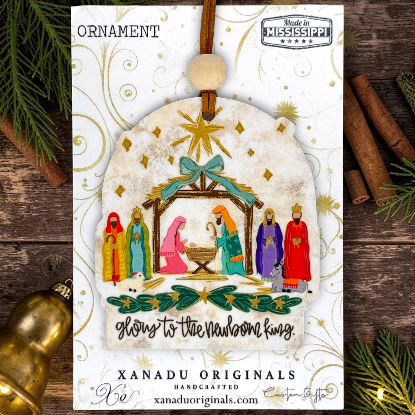 A handcrafted Christmas ornament by Xanadu Originals, featuring a nativity scene illustration on a white background with gold swirls. The ornament is shown hanging from a brown string with a wooden bead, surrounded by festive props like pine branches, cinnamon sticks, and a gold bell on a dark wood surface.