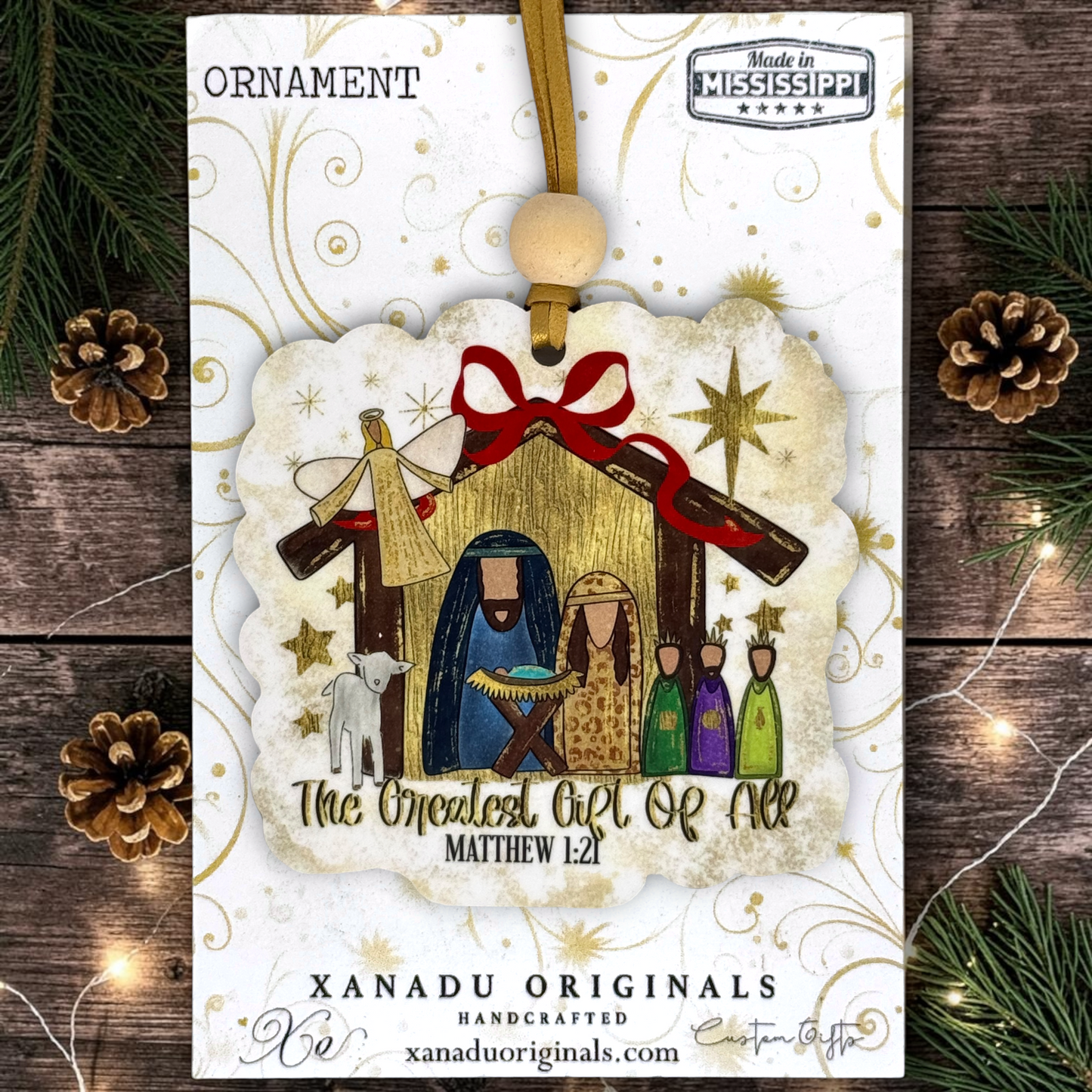 A Christmas ornament package from Xanadu Originals featuring a nativity scene ornament with Mary, Joseph, baby Jesus, the angel, and the three wise men, hanging from a wooden bead on a white card with gold swirls, surrounded by pine branches and pinecones.