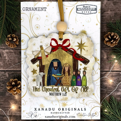 A Christmas ornament package from Xanadu Originals featuring a nativity scene ornament with Mary, Joseph, baby Jesus, the angel, and the three wise men, hanging from a wooden bead on a white card with gold swirls, surrounded by pine branches and pinecones.