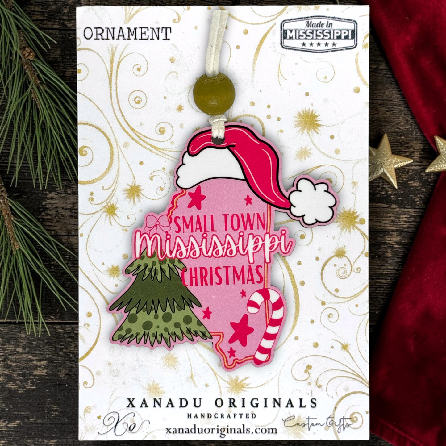 A Christmas ornament on a card featuring a layered ornament design with a pink state-of-Mississippi shape, a red Santa hat, a green tree, and a candy cane, with the phrase “Small Town Mississippi Christmas.” The card is branded Xanadu Originals and shows “Made in Mississippi.