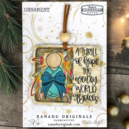 Handcrafted Christmas ornament card by Xanadu Originals, featuring a stylized angel ornament with a turquoise bow, colorful star accents, and the phrase 'A THRILL OR HOPE THE WEARY WORLD ENJOYS.' The card is displayed on a rustic wood surface with pine branches and gold ornaments around it.
