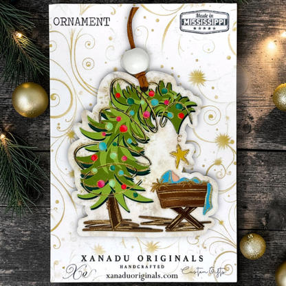 A Christmas ornament card featuring a handcrafted ornament design of a green Christmas tree with colorful ornaments and a wooden baby crib. The card is branded 'XANADU ORIGINALS' and shows 'Made in Mississippi' at the top, set against a rustic dark wood background with pine needles and gold ornaments.